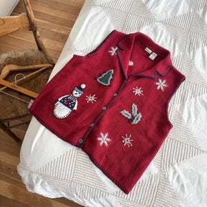 May include: Red sleeveless sweater vest with a Christmas theme. The vest has embroidered snowflakes, a snowman, a Christmas tree, and holly leaves. The vest has a button-up front.