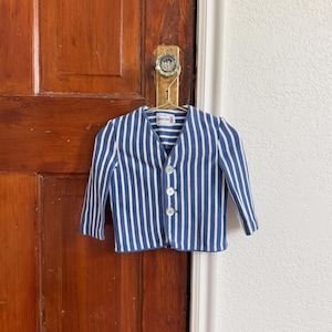 May include: A blue and white striped jacket with three white buttons. The jacket is hanging on a gold hanger from a door knob. The jacket has a V-neck and long sleeves. The label reads "sunny day".