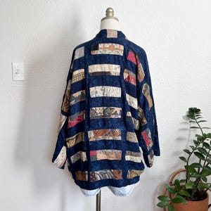 Puede incluir: Chaqueta patchwork azul oscuro con paneles rectangulares de tela en varios patrones y colores. La chaqueta se exhibe en un maniqu&iacute;, mostrando su dise&ntilde;o y textura &uacute;nicos. La chaqueta tiene un corte holgado y mangas largas.