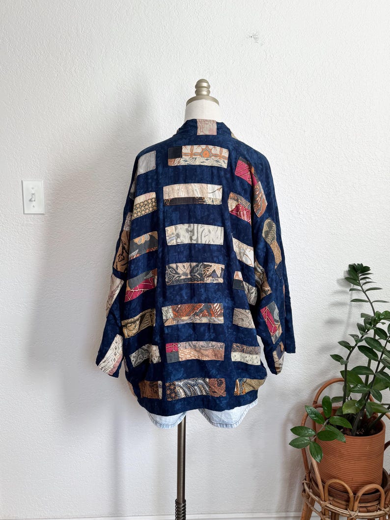 Puede incluir: Chaqueta patchwork azul oscuro con paneles rectangulares de tela en varios patrones y colores. La chaqueta se exhibe en un maniqu&iacute;, mostrando su dise&ntilde;o y textura &uacute;nicos. La chaqueta tiene un corte holgado y mangas largas.