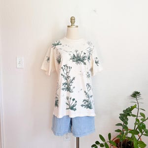 K&ouml;nnte beinhalten: Cremefarbenes T-Shirt mit einem botanischen Druck von gr&uuml;nen Pflanzen und Blumen. Das T-Shirt wird mit hellblauen Jeansshorts kombiniert. Das Outfit wird auf einer Schaufenquin vor einer wei&szlig;en Wand pr&auml;sentiert.