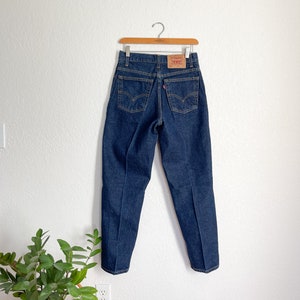 May include: A pair of dark blue denim jeans hanging on a wooden hanger against a white wall. The jeans have a classic five-pocket style and a straight leg.