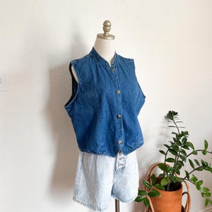 May include: A vintage denim vest with a mandarin collar and button-up front. The vest is a medium wash blue and is paired with light wash denim shorts. The outfit is displayed on a mannequin against a white wall.