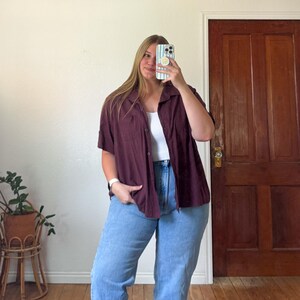 May include: A short-sleeved, button-down shirt in a deep maroon color with a collar and a chest pocket, worn over a white ribbed tank top paired with light blue denim jeans. The shirt features a textured fabric and a relaxed, loose-fitting cut.