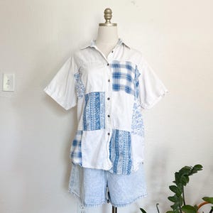 May include: A white patchwork button-down shirt with blue plaid and floral patterned panels. The shirt is paired with light blue denim shorts. The outfit is displayed on a mannequin.