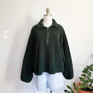 May include: A dark green fleece pullover with a half-zip closure and a collared neckline. The plush, textured fabric is displayed on a mannequin, paired with light blue denim shorts. The garment is a cozy, casual top.