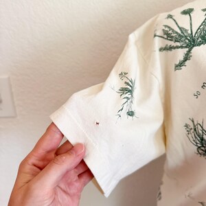 K&ouml;nnte beinhalten: Cremefarbenes Kurzarm-T-Shirt mit gr&uuml;nem botanischem Druck. Das Design zeigt verschiedene Pflanzenillustrationen. Der &Auml;rmel ist sichtbar und zeigt den Saum und die Textur des Stoffes.