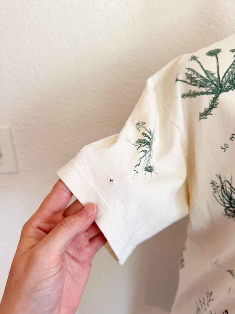 K&ouml;nnte beinhalten: Cremefarbenes Kurzarm-T-Shirt mit gr&uuml;nem botanischem Druck. Das Design zeigt verschiedene Pflanzenillustrationen. Der &Auml;rmel ist sichtbar und zeigt den Saum und die Textur des Stoffes.