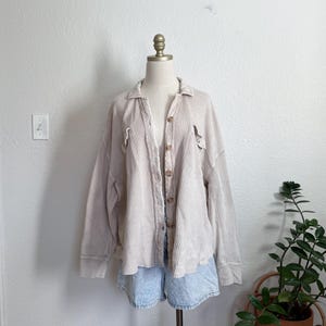 May include: A light beige waffle-knit shirt with long sleeves and a button-up front. The shirt is displayed on a mannequin with light blue denim shorts. The shirt has two front pockets and a collar. The background is a plain white wall.