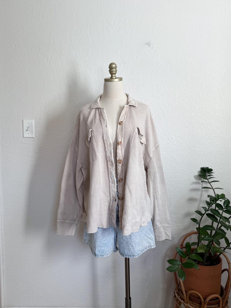 May include: A light beige waffle-knit shirt with long sleeves and a button-up front. The shirt is displayed on a mannequin with light blue denim shorts. The shirt has two front pockets and a collar. The background is a plain white wall.