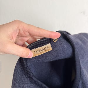 May include: A navy blue garment with a dark grey trim is shown in close-up. The label reads "LANDHAUS CAA NATURALLY". The fabric appears soft and textured. The garment's design is simple and elegant.