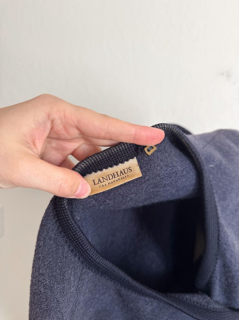 May include: A navy blue garment with a dark grey trim is shown in close-up. The label reads "LANDHAUS CAA NATURALLY". The fabric appears soft and textured. The garment's design is simple and elegant.