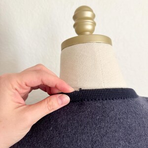 May include: A dark grey garment with a black trim around the neckline is displayed on a mannequin. A hand is touching the trim. The mannequin has a gold-coloured top.