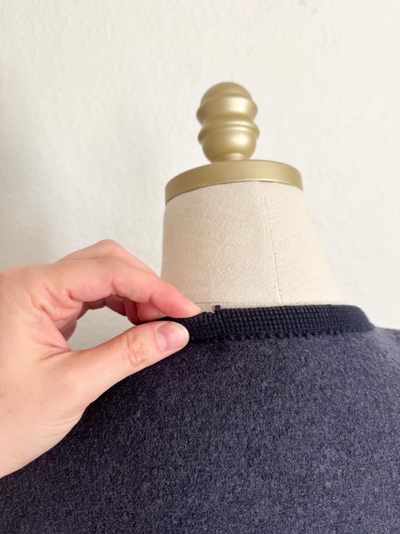 May include: A dark grey garment with a black trim around the neckline is displayed on a mannequin. A hand is touching the trim. The mannequin has a gold-coloured top.