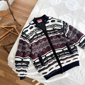 Peut inclure: Un pull noir &agrave; fermeture &eacute;clair avec un motif g&eacute;om&eacute;trique color&eacute; dans les tons de rouge, blanc, vert et noir. Le pull a un col et des poignets c&ocirc;tel&eacute;s.