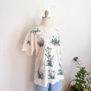 K&ouml;nnte beinhalten: Cremefarbenes T-Shirt mit einem botanischen Druck verschiedener Pflanzen in Gr&uuml;nt&ouml;nen. Das Shirt hat kurze &Auml;rmel und einen Rundhalsausschnitt. Der Text "so many weeds, so little time" ist auf dem Shirt aufgedruckt. Dazu werden hellblaue Jeansshorts getragen.