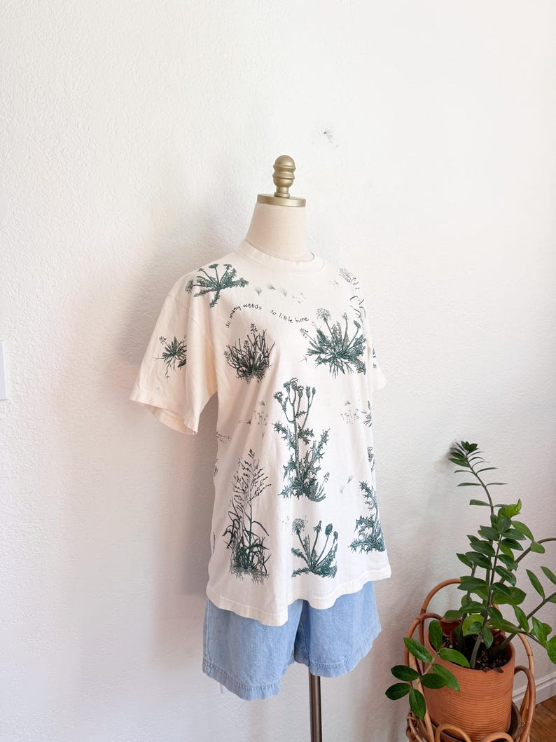 K&ouml;nnte beinhalten: Cremefarbenes T-Shirt mit einem botanischen Druck verschiedener Pflanzen in Gr&uuml;nt&ouml;nen. Das Shirt hat kurze &Auml;rmel und einen Rundhalsausschnitt. Der Text "so many weeds, so little time" ist auf dem Shirt aufgedruckt. Dazu werden hellblaue Jeansshorts getragen.