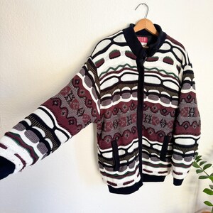 Peut inclure: Un cardigan vintage &agrave; fermeture &eacute;clair avec un motif g&eacute;om&eacute;trique color&eacute; dans les tons de rouge, blanc, noir et vert. Le cardigan a deux poches sur le devant.