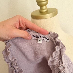 May include: A light purple top with ruffled edges and the text "Sweet Mariah" on a tag. The top is size small and appears to be made of a soft, textured fabric. The top is displayed on a mannequin.