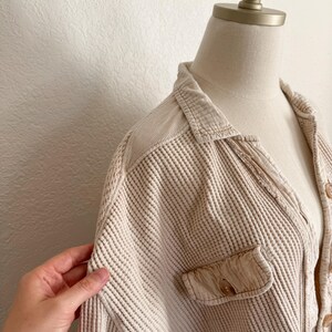 May include: Beige waffle-knit shirt with a collared neckline and a single chest pocket. The shirt is buttoned and displayed on a mannequin. The fabric has a textured, grid-like pattern.