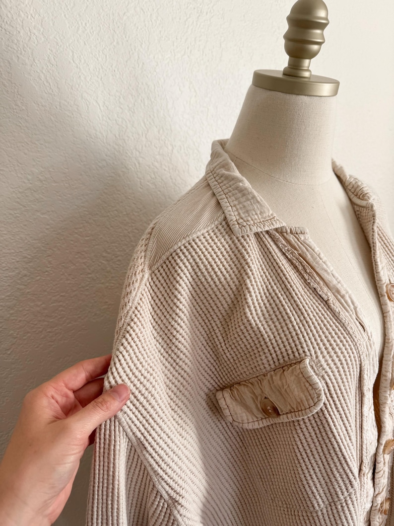 May include: Beige waffle-knit shirt with a collared neckline and a single chest pocket. The shirt is buttoned and displayed on a mannequin. The fabric has a textured, grid-like pattern.