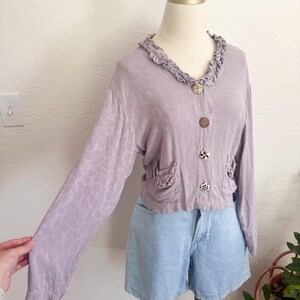May include: A light purple, long-sleeved cardigan with decorative buttons and ruffled collar. The cardigan has two small pockets and is paired with light blue denim shorts. The fabric has a subtle floral pattern.