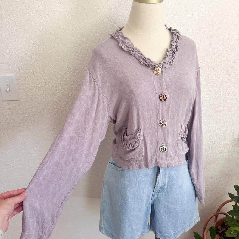 May include: A light purple, long-sleeved cardigan with decorative buttons and ruffled collar. The cardigan has two small pockets and is paired with light blue denim shorts. The fabric has a subtle floral pattern.