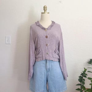 May include: A light purple cardigan with long sleeves, a ruffled collar, and decorative buttons. The cardigan has two small pockets with ruffled edges and is paired with light blue denim shorts. The outfit is displayed on a mannequin.
