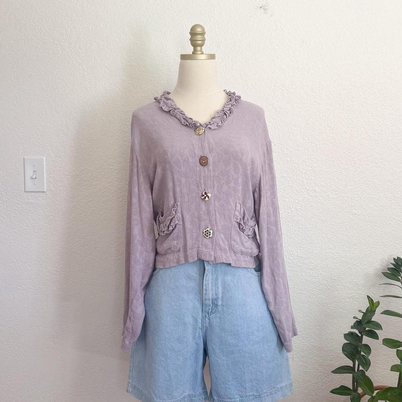 May include: A light purple cardigan with long sleeves, a ruffled collar, and decorative buttons. The cardigan has two small pockets with ruffled edges and is paired with light blue denim shorts. The outfit is displayed on a mannequin.