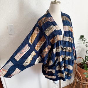 Puede incluir: Chaqueta estilo kimono azul marino con dise&ntilde;o patchwork. La chaqueta presenta parches rectangulares en varios colores y patrones, incluyendo beige, marr&oacute;n y rosa. La prenda se exhibe en un maniqu&iacute;, mostrando su ajuste holgado y frente abierto.