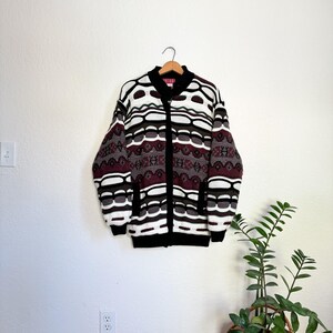 Peut inclure: Un cardigan &agrave; motif blanc, noir, marron et bordeaux avec une fermeture &eacute;clair noire. Le cardigan est suspendu &agrave; un cintre contre un mur blanc.
