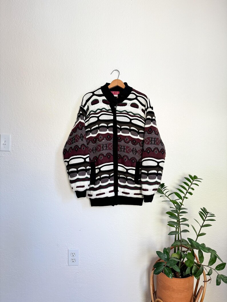 Peut inclure: Un cardigan &agrave; motif blanc, noir, marron et bordeaux avec une fermeture &eacute;clair noire. Le cardigan est suspendu &agrave; un cintre contre un mur blanc.