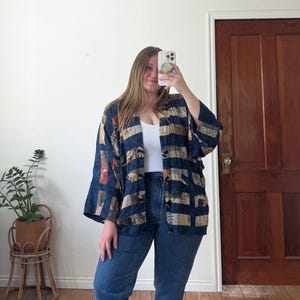 Puede incluir: Chaqueta estampada en azul marino, beige y rojo con frente abierto. La chaqueta tiene un corte holgado y mangas anchas. La persona lleva vaqueros azules y una camiseta blanca. En el fondo, una puerta de madera y una planta en una maceta de mimbre.
