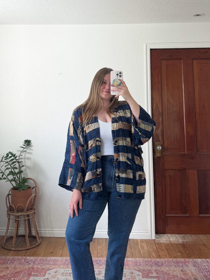 Puede incluir: Chaqueta estampada en azul marino, beige y rojo con frente abierto. La chaqueta tiene un corte holgado y mangas anchas. La persona lleva vaqueros azules y una camiseta blanca. En el fondo, una puerta de madera y una planta en una maceta de mimbre.