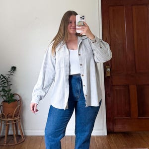 May include: A light-colored, vertically striped button-down shirt with long sleeves and a white tank top. The shirt is paired with blue jeans. The outfit is casual and comfortable.