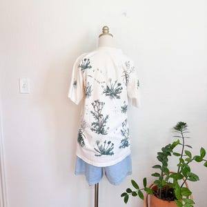 K&ouml;nnte beinhalten: Ein wei&szlig;es T-Shirt mit einem botanischen Druck von gr&uuml;nen Pflanzen und dem Text "weeds to live here". Das T-Shirt wird mit hellblauen Jeansshorts kombiniert. Das Outfit wird auf einer Schaufensterpuppe vor einer wei&szlig;en Wand pr&auml;sentiert.