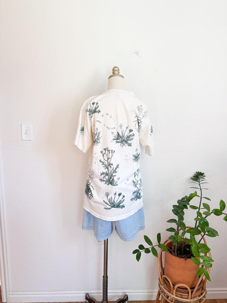 K&ouml;nnte beinhalten: Ein wei&szlig;es T-Shirt mit einem botanischen Druck von gr&uuml;nen Pflanzen und dem Text "weeds to live here". Das T-Shirt wird mit hellblauen Jeansshorts kombiniert. Das Outfit wird auf einer Schaufensterpuppe vor einer wei&szlig;en Wand pr&auml;sentiert.