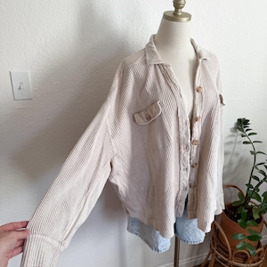 May include: Beige waffle-knit shirt with long sleeves, a collared neckline, and button-up front. The shirt features two front pockets and is displayed on a mannequin. The sleeves are cuffed at the wrists.