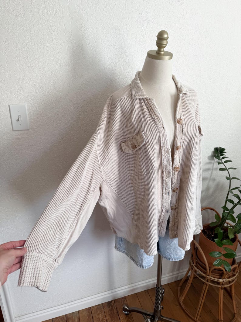 May include: Beige waffle-knit shirt with long sleeves, a collared neckline, and button-up front. The shirt features two front pockets and is displayed on a mannequin. The sleeves are cuffed at the wrists.