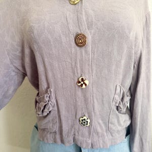 May include: A light purple jacket with floral embossed fabric, featuring decorative buttons and ruffled pockets. The buttons have unique designs, including a spiral and flower shapes. The jacket is cropped and has long sleeves.