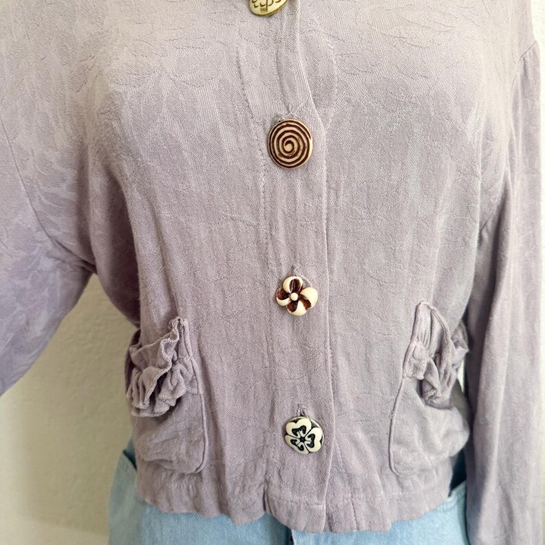 May include: A light purple jacket with floral embossed fabric, featuring decorative buttons and ruffled pockets. The buttons have unique designs, including a spiral and flower shapes. The jacket is cropped and has long sleeves.