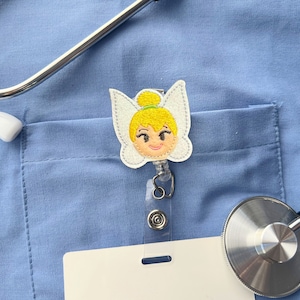 May include: A white and yellow embroidered badge featuring Tinkerbell, a Disney fairy, with a retractable badge reel attached. The badge is on a blue scrub top with a stethoscope.