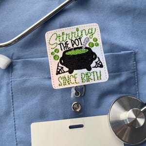 May include: A white badge reel with the text "Stirring the Pot Since Birth" and a black cauldron design with green accents. The badge reel is attached to a light blue shirt, with a stethoscope in the background.