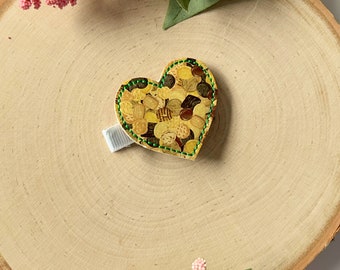 May include: A heart-shaped hair clip with a gold background and a green stitched border. The clip features a pattern of various cookies and candies.
