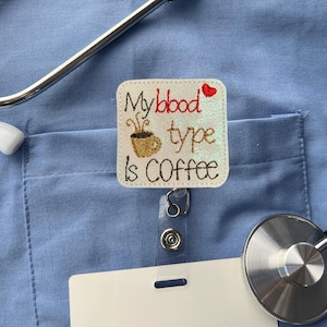 May include: A square badge reel with the text "My blood type is COFFEE" and a coffee cup graphic. The badge is white with glitter accents and a red heart. A stethoscope and blank name tag are also visible.