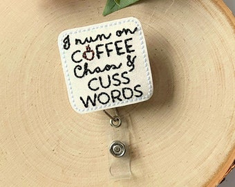 May include: White square badge holder with a black embroidered design that reads "I run on coffee, chaos & cuss words". A coffee cup icon is included in the design. The badge holder is attached to a clear plastic retractable badge reel.