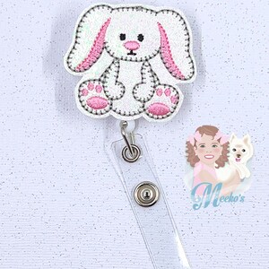 May include: White glitter bunny with pink ears and paws, sitting on a clear plastic badge holder with silver clips. The badge holder is attached to a white background with glitter.