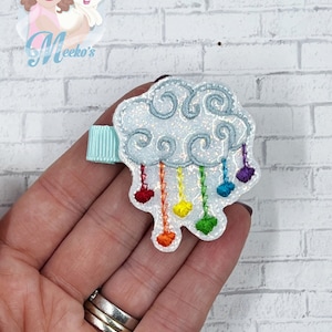 Op de afbeelding: Een lichtblauwe glitterwolk met een regenboog van harten die eraan hangen. De wolk is bevestigd aan een lichtblauwe alligatorclip.