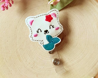 May include: White felt mermaid with a pink flower on her head and a teal tail. The mermaid is attached to a clear plastic retractable badge holder.