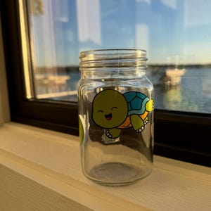 Puede incluir: Un tarro de cristal transparente con tapa de rosca. Una pegatina de tortuga de dibujos animados con caparazón verde, detalles azules y una cara sonriente está en el tarro. El tarro está sobre un alféizar blanco con vistas a un cuerpo de agua y el cielo.